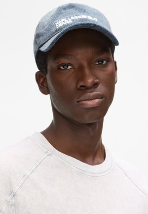 Νέος Μαύρος άνδρας φοράει ανοιχτό μπλε καπέλο από τζιν με λευκό κείμενο και λευκό μπλουζάκι με στρογγυλή λαιμόκοψη, κοιτάζει μπροστά με ουδέτερη έκφραση.