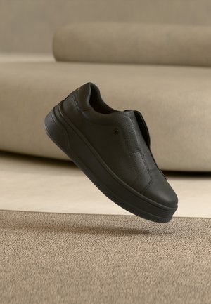Μαύρο δερμάτινο παπούτσι slip-on που αιωρείται πάνω από ένα μπεζ χαλί με μια καμπυλωτή μπεζ καναπέ στο φόντο.
