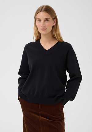 Femme aux cheveux blonds lisses portant un pull noir col V et une jupe en velours marron sur un fond clair uni.