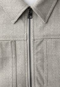 Primer plano de una chaqueta de tela gris de espiga con cremallera metálica negra y cuello puntiagudo.