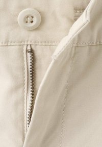 Nahaufnahme eines beigen Baumwollstoffs mit einem silbernen Metallreißverschluss und einem weißen Knopf mit eingraviertem Text, sichtbare Nähte.