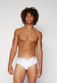 Weiße Baumwollslips mit einem schwarzen Lacoste-Logo am Bund, mit glatter Textur und figurbetontem Design, getragen von einem Modell.