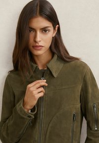 Õlivärvi rohelise seemisnahast jakk kraega, esiküljel tõmblukk ja kaks nurga all asetsevat tõmblukuga taskut, millel on kontrastsed mustad riistvara detailid.