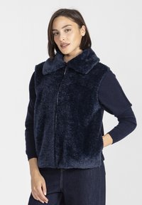 Kobieta ubrana w granatowy, puszysty kamizelkę zapinaną na zamek z szerokim kołnierzem na długim rękawie granatowej koszuli i ciemnych dżinsach, pozująca z jedną ręką w kieszeni.