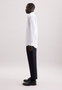 Camicia bianca a maniche lunghe con colletto a bottoni e taschino sul petto, abbinata a pantaloni scuri su misura e scarpe nere, vista di lato.