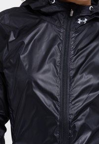 Czarna wodoodporna kurtka z zamkiem błyskawicznym, lekka, błyszcząca tkanina, regulowany kaptur oraz logo Under Armour z przodu.