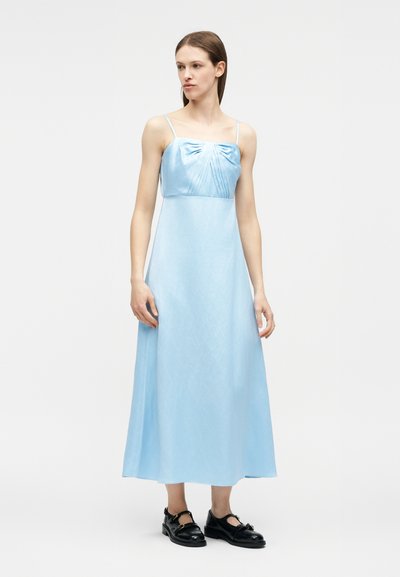 Γυναίκα που φοράει αμάνικο midi φόρεμα ανοιχτού γαλάζιου χρώματος με πιετέ μπούστο και μαύρα κλειστά παπούτσια, στέκεται σε απλό λευκό φόντο.