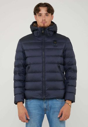 Junger Mann trägt eine marineblaue Steppjacke mit Kapuze und blaue Jeans, steht vor einem einfarbigen hellgrauen Hintergrund.