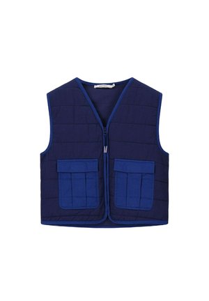 Jongen staat met handen in de zakken, draagt een paarse trui en korte broek, een blauw doorgestikt vest, witte sokken en groene sneakers.
