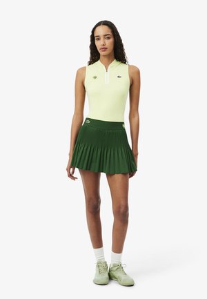 Femme debout portant un haut sans manches jaune zippé, une jupe plissée vert foncé, des chaussettes blanches et des baskets vert clair sur fond blanc.