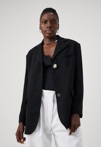 Blazer negru cu o croială lejeră, închidere cu un singur nasture și o broșă argintie. Țesătura este netedă; are un guler cu deschidere pe față și două buzunare frontale.