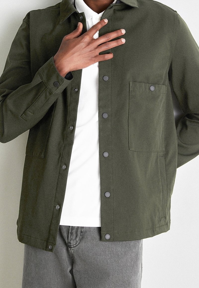 Persoon draagt een olijfgroene jas met knopen over een wit shirt en grijze broek, met één hand die de borst nabij de kraag raakt.