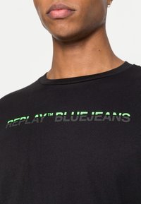 Camiseta de algodón negra con cuello redondo y un estampado gráfico en verde y gris que dice "REPLAY™ BLUEJEANS" en el pecho.