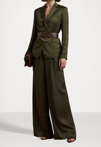Olivgrön skräddarsydd kavaj med bälte som har en guldkedja. Byxor med vida ben i matchande tyg, texturerade med en slät finish.