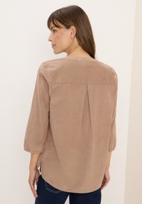 Femme aux cheveux bruns portant un blouse loose, de couleur marron clair, avec des manches trois-quarts et un jean bleu foncé, tournée de dos à la caméra.