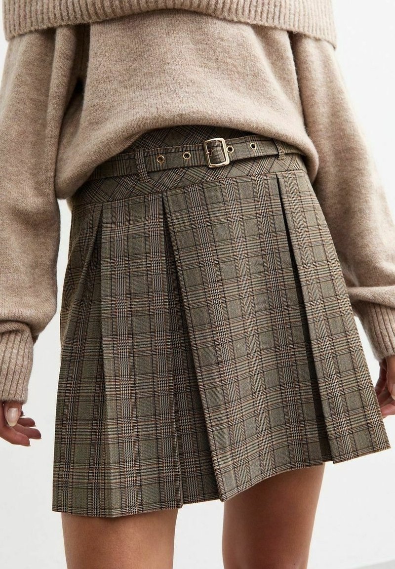 Femme portant une jupe plissée à carreaux marron avec une ceinture assortie et un pull en tricot beige, montrant la taille et les mains.