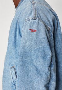 Ανοιχτό μπλε τζιν μπουφάν με ριχτό γιακά, πλαϊνές τσέπες, εμφανή ραφή και μικρό κόκκινο κεντητό λογότυπο στο αριστερό μανίκι.