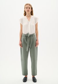 Blusa bianca a maniche corte con scollo arricciato, abbinata a pantaloni a vita alta verde chiaro con cintura in tessuto e cuciture laterali. Scarpe nere slip-on.