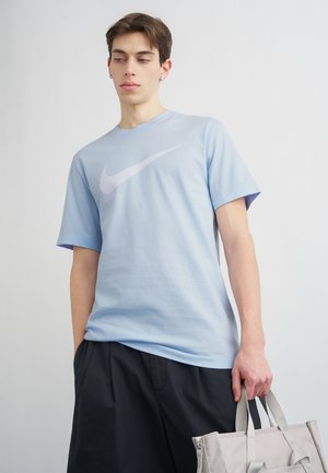 Joven con camiseta azul claro de Nike sosteniendo una bolsa tote beige, mirando hacia abajo y de lado, sobre un fondo liso.