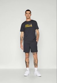 Férfi áll egy szürke háttér előtt, fekete New Balance pólóban, fekete rövidnadrágban, fehér zoknikban, fehér sportcipőben, és tetoválásokat mutat a karjain és lábain.