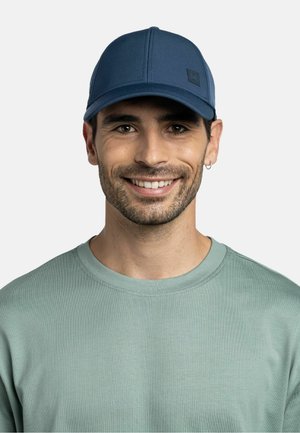 Smiling man wearing a blue cap and light green crew-neck shirt, with short dark hair and a small hoop earring on a light background.