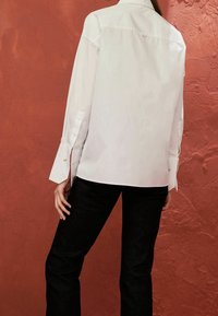 Chemise blanche à manches longues avec un col boutonné, ornée de détails aux poignets et une coupe droite, associée à un pantalon en denim foncé.