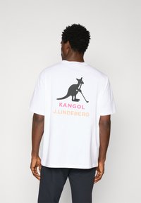 Hvid bomulds t-shirt med et sort kenguru-grafik, der holder en golfkølle, samt "KANGOL" i pink og "J. LINDEBERG" i orange tekst.