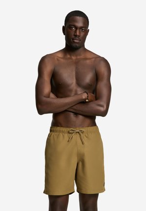 Muskelbepackter Mann mit verschränkten Armen, der senffarbene Badehosen und ein schwarzes Perlenarmband trägt, steht vor einem schlichten weißen Hintergrund.
