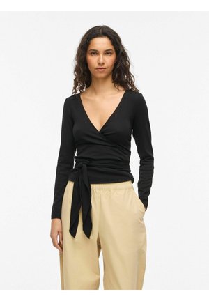 Kobieta o kręconych ciemnych włosach nosi czarną bluzkę wiązaną w talii z długim rękawem oraz beżowe luźne spodnie, stojąc na białym tle.