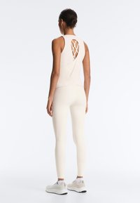 Woman wearing beige sleeveless top with crisscross back cutout and matching leggings, standing against a light background.