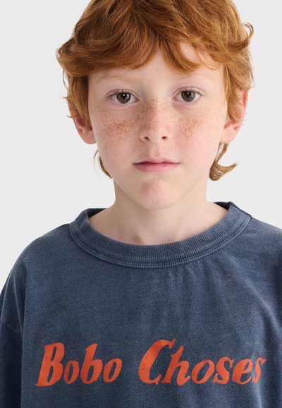Bambino rosso dai capelli rossi con lentiggini indossa una maglietta blu scuro con la scritta arancione "Bobo Choses", guarda dritto davanti a uno sfondo semplice.