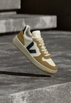 Baskets blanches et beige avec logo noir flottant au-dessus des marches en béton gris en extérieur.