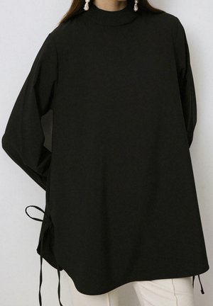 Femme portant une tunique noire ample à manches longues avec des liens sur les côtés et des boucles d'oreilles en perles tombantes, associée à un pantalon de couleur claire.