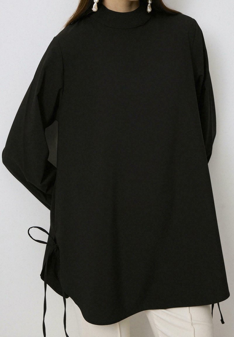 Femme portant une tunique noire ample à manches longues avec des liens sur les côtés et des boucles d'oreilles en perles tombantes, associée à un pantalon de couleur claire.