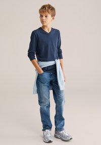 Tengerészkék hosszú ujjú V-nyakú ing, világoskék pulóver a derék körül megkötve, kék farmernadrág és szürke tornacipő. Egyszerű dizájn, hétköznapi stílus.