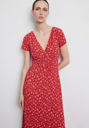 Donna che indossa un vestito rosso a maniche corte con piccola stampa floreale bianca e dettaglio a nodo attorcigliato in vita, in piedi contro un muro semplice.