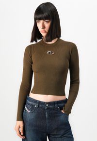 Kobieta z prostymi ciemnymi włosami, ubrana w oliwkową, prążkowaną bluzkę typu crop top z długim rękawem i wycięciem na klatce piersiowej oraz ciemnoniebieskie spodnie jeansowe z wysokim stanem.