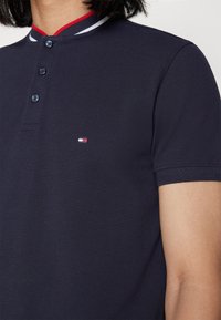 Närbild av en person som bär en marinblå kortärmad polo med tre knappar och en liten röd, vit och blå flagglogotyp på bröstet.