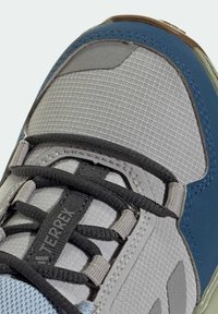 Grå syntetisk vandresko med meshstof, sorte snørebånd og en blå detalje nær anklen. Har forstærket tåhætter og struktureret overdel.