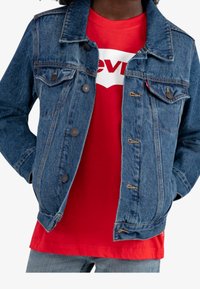 Chaqueta de mezclilla con acabado azul, bolsillos en el pecho y cierres de botón, llevada sobre una camiseta roja con texto blanco y un logo.