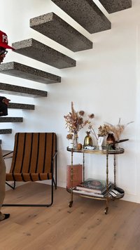 Crni metalni stolac s smešastim i crnim prugastim materijalom, okrugli zlatni metalni kolica s vazama, suvim cvećem i knjigama pored betonskih stepenica.