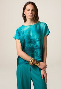 Blusa di seta turchese con un motivo decorativo ispirato alla natura, maniche corte e vestibilità rilassata, abbinata a pantaloni larghi teal coordinati e braccialetti dorati.