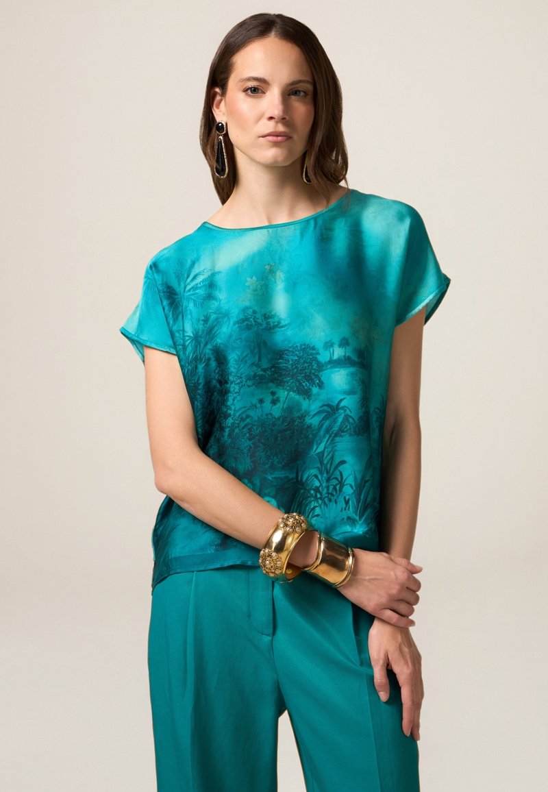 Blusa di seta turchese con un motivo decorativo ispirato alla natura, maniche corte e vestibilità rilassata, abbinata a pantaloni larghi teal coordinati e braccialetti dorati.