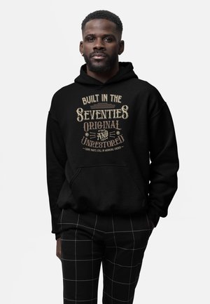 Homme portant un sweat à capuche noir avec le texte "Built in the Seventies Original and Unrestored" et un pantalon noir à motif quadrillé blanc, debout devant un fond blanc.