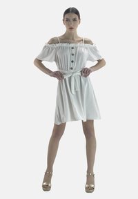 Vestido blanco sin hombros con mangas cortas abullonadas, detalles de botones y cintura ceñida, con un nudo en la parte delantera y falda fluida. Tacones de plataforma dorados.