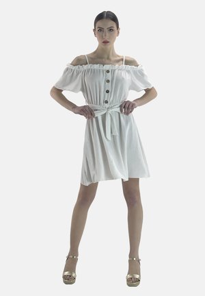 Weißes schulterfreies Kleid mit kurzen Puffärmeln, Knopfdetails und taillierter Taille, ausgestattet mit einem Frontbindeband und einem fließenden Rock. Goldene Plateauschuhe.