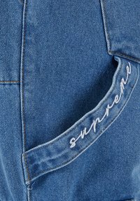 Blauer Denim-Stoff mit leicht verblichenem Muster. Verfügt über eine auffällige Tasche mit "supreme" in Weiß auf dem Rand gestickt.