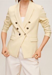 Blazer su misura color crema con tasche a patta doppiopetto e bottoni neri lucidi. Tessuto leggero con spalle strutturate.