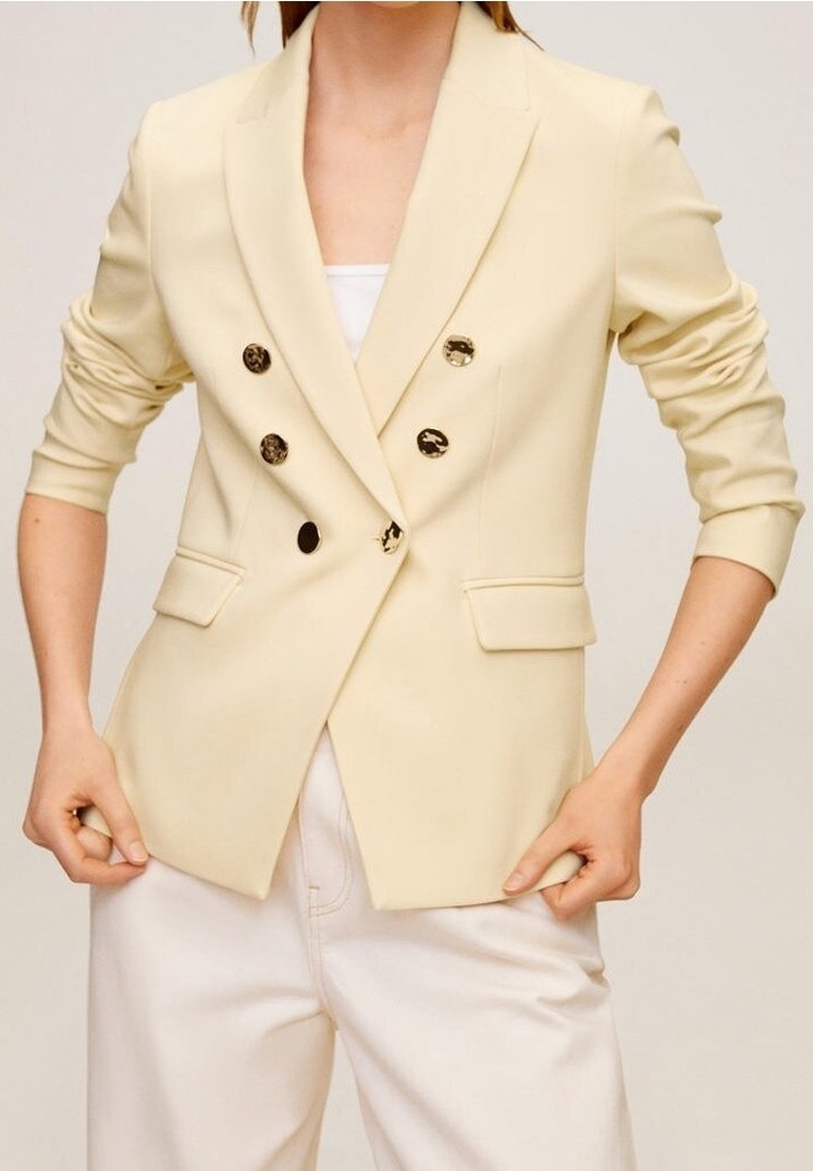 Blazer su misura color crema con tasche a patta doppiopetto e bottoni neri lucidi. Tessuto leggero con spalle strutturate.