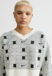Adīta balta džemperis ar melnu ģeometrisku rakstu, v-veida kakla dizains, ribota apakšmalas, ar maziem melniem Nike logo visā virsmas.